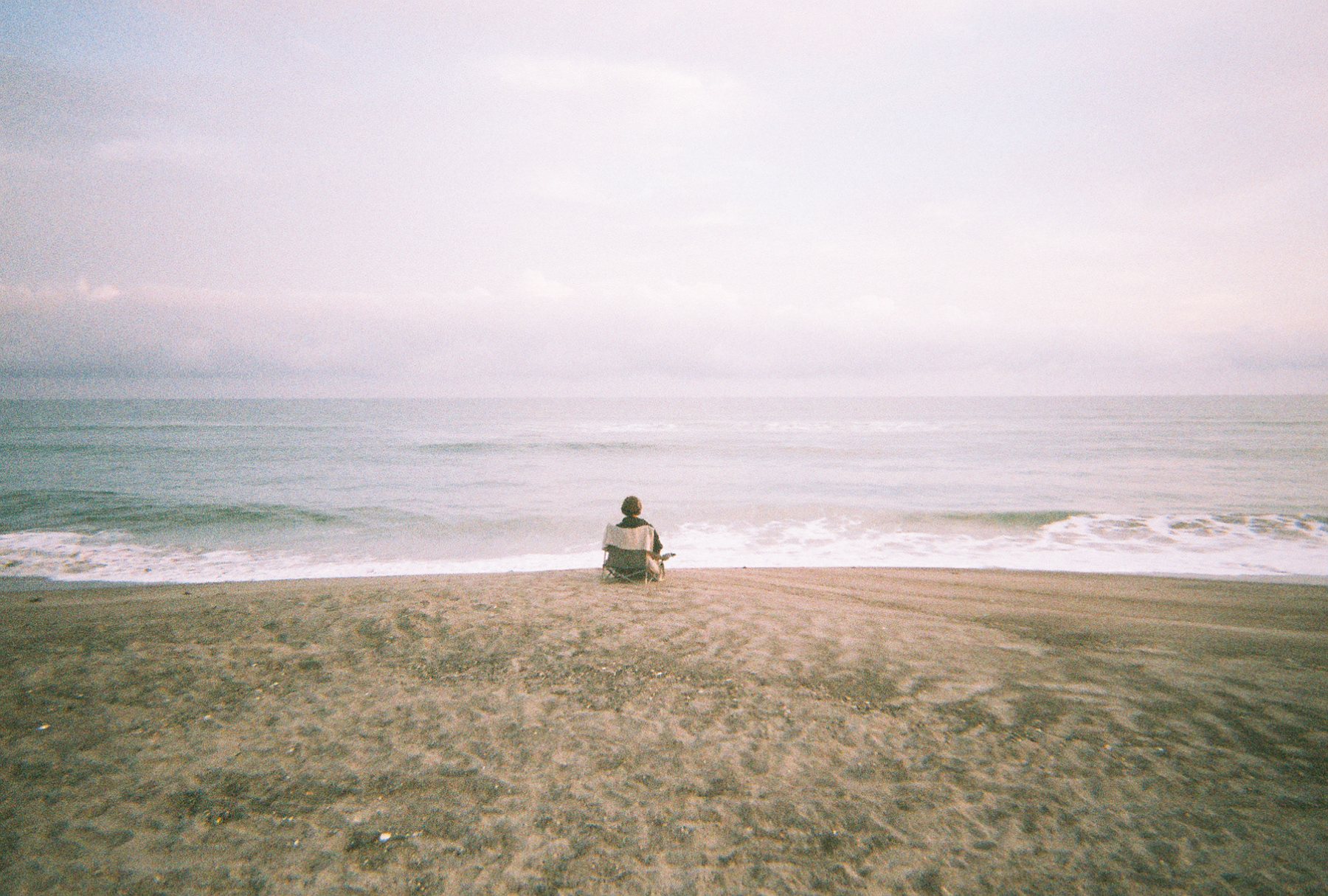 A figure sitting in a chair facing the ocean on an empty beach