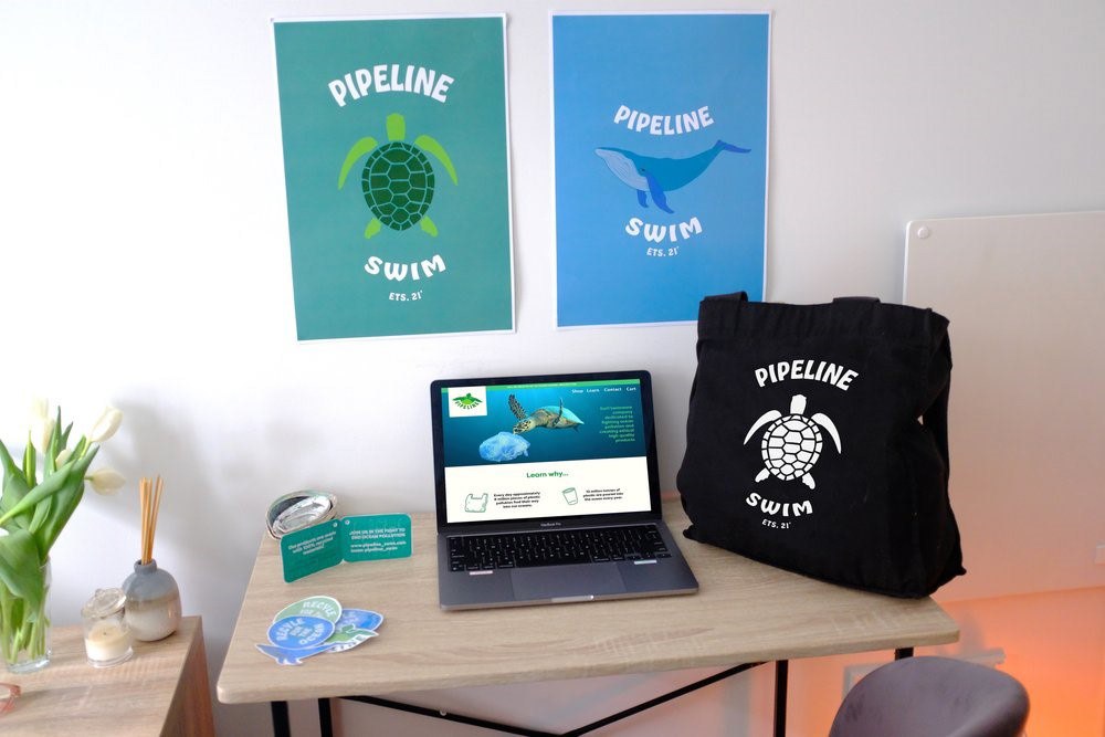 Pipeline Swim posters, tote bag, stickers, and website mockup displayed on a desk